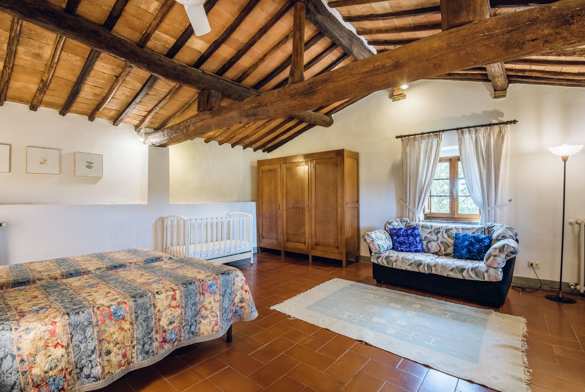 A room with a wooden ceiling, a bed with a floral bedspread, a sofa with patterned cushions, a radiator, and a window with curtains.