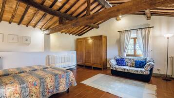 A room with a wooden ceiling, a bed with a floral bedspread, a sofa with patterned cushions, a radiator, and a window with curtains.