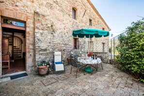Une terrasse extérieure avec un bâtiment en pierre, une table dressée pour le repas et un parasol vert.