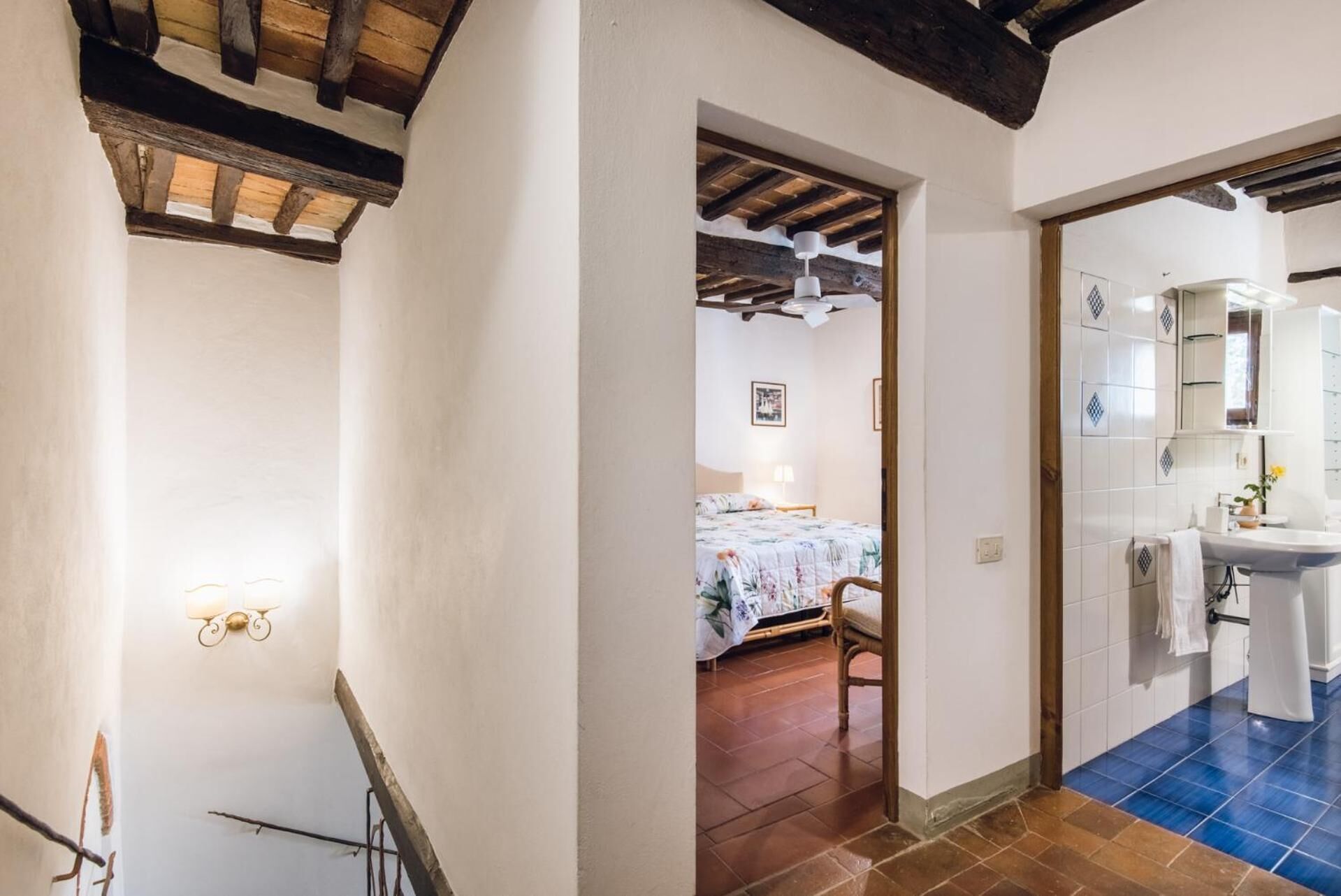 A bedroom with a bed, a chair, and a nightstand. There is a bathroom with a sink and a mirror. The room has wooden beams on the ceiling and tiled floors.