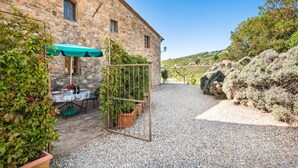 Un edificio in pietra con un vialetto sterrato che conduce a un tavolo apparecchiato per mangiare, immerso nel verde e con un vigneto sullo sfondo.