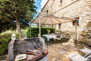 Une terrasse extérieure avec un barbecue, une table recouverte d’une nappe blanche et, en arrière-plan, un bâtiment en pierre.