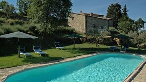 Una piscina con acqua cristallina, circondata da sdraio e ombrelloni, con sullo sfondo un edificio in pietra e un giardino.