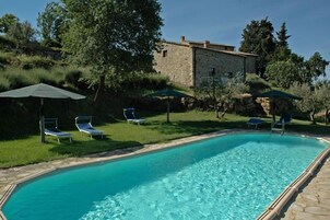 Una piscina con acqua cristallina, circondata da sdraio e ombrelloni, con sullo sfondo un edificio in pietra e un giardino.