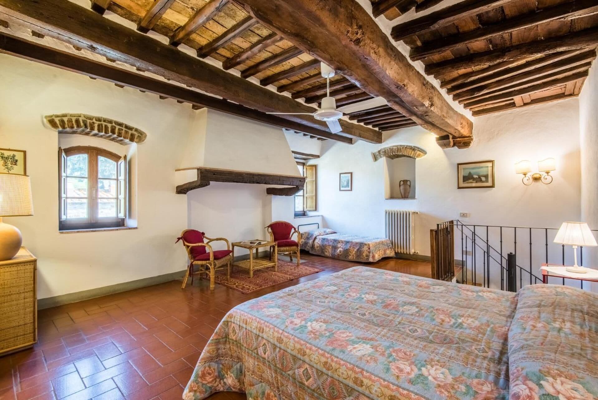Quarto rústico com vigas de madeira aparentes, cama com roupa de cama florida, cadeiras de vime e uma pequena mesa.