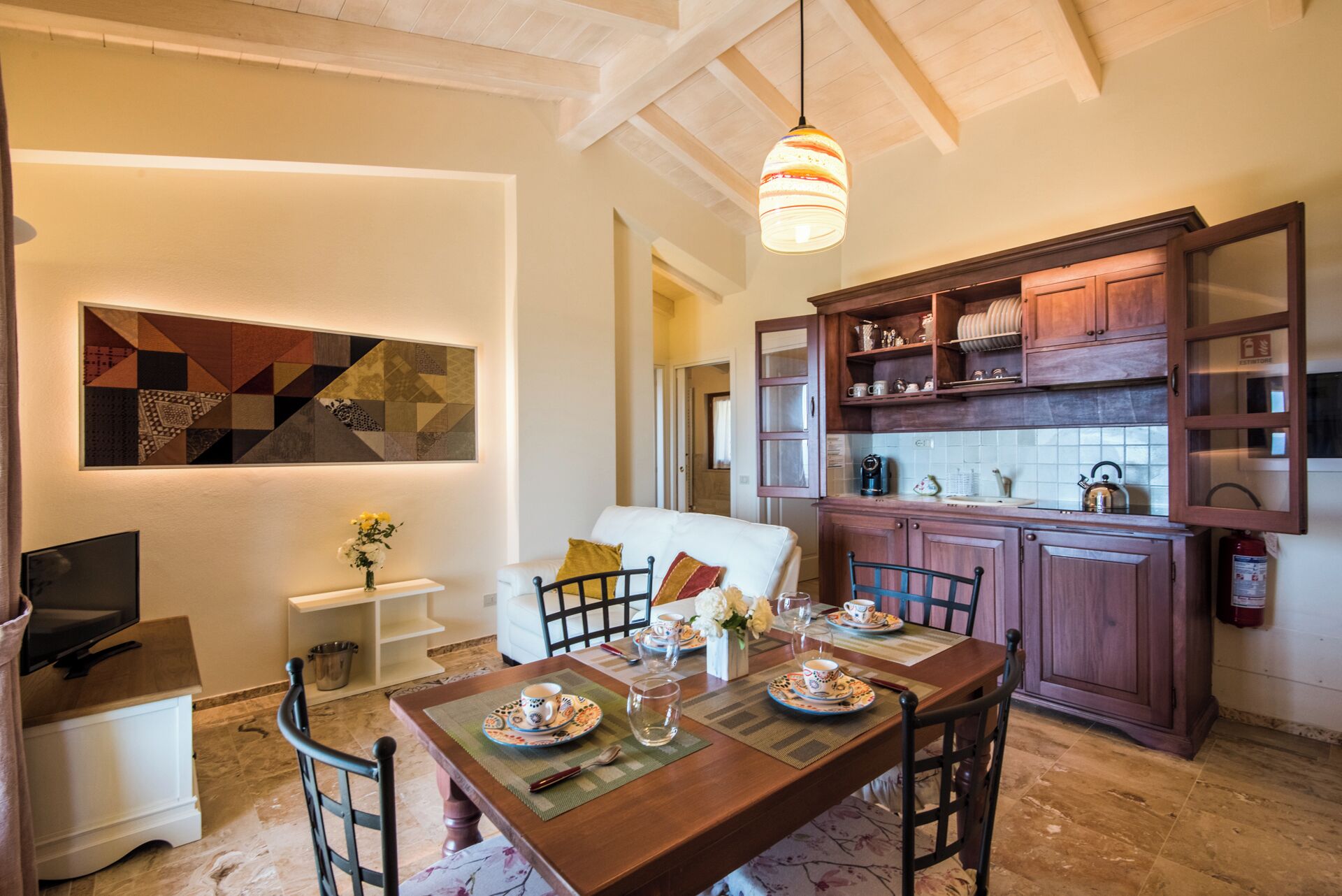 On aperçoit un coin repas avec une table en bois dressée pour quatre personnes, entourée de chaises. Au fond, on distingue une cuisine avec des meubles en bois et un dosseret carrelé.