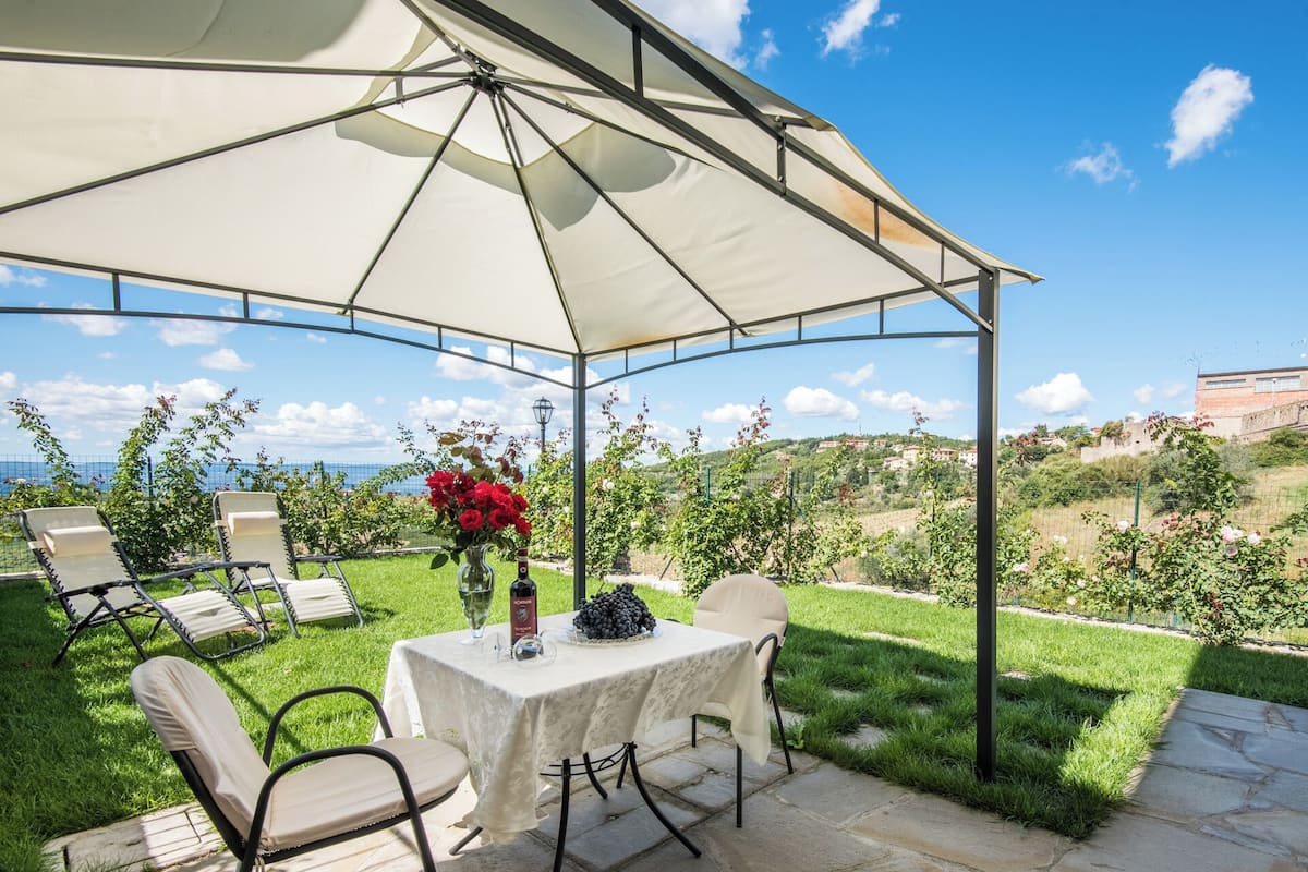 Une terrasse extérieure avec une table et des chaises, abritée par un auvent, d’où l’on peut admirer un jardin luxuriant et un paysage lointain.