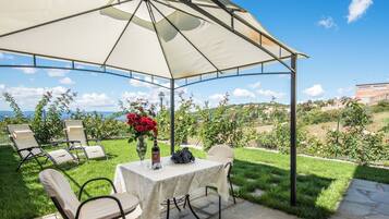 Une terrasse extérieure avec une table et des chaises, abritée par un auvent, d’où l’on peut admirer un jardin luxuriant et un paysage lointain.