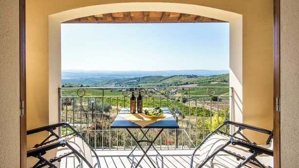 A balcony with a table, chairs, and a bottle of wine, overlooking a vineyard and distant hills. - Casa Vacanze Squarcialupi (Castellina in Chianti)