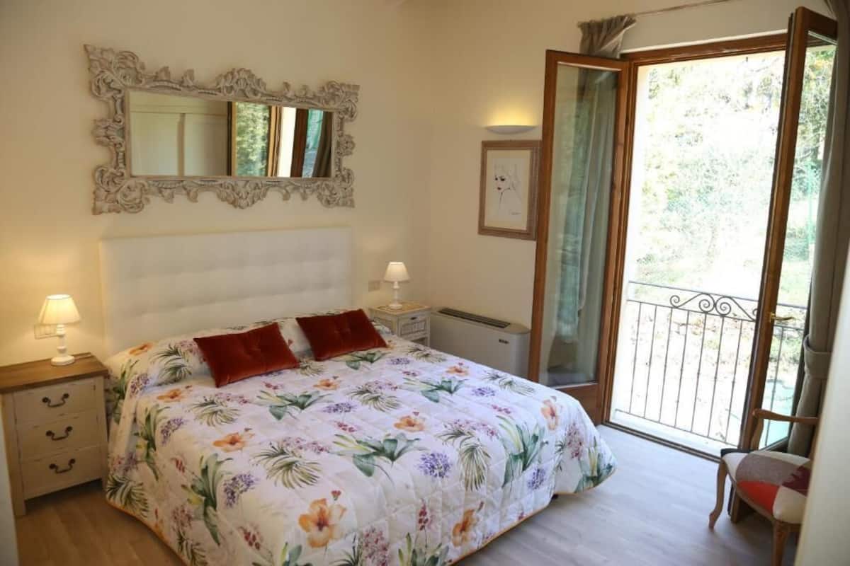 Une chambre à coucher comprenant un lit, une table de chevet, une commode, un miroir et un balcon donnant sur un paysage verdoyant.