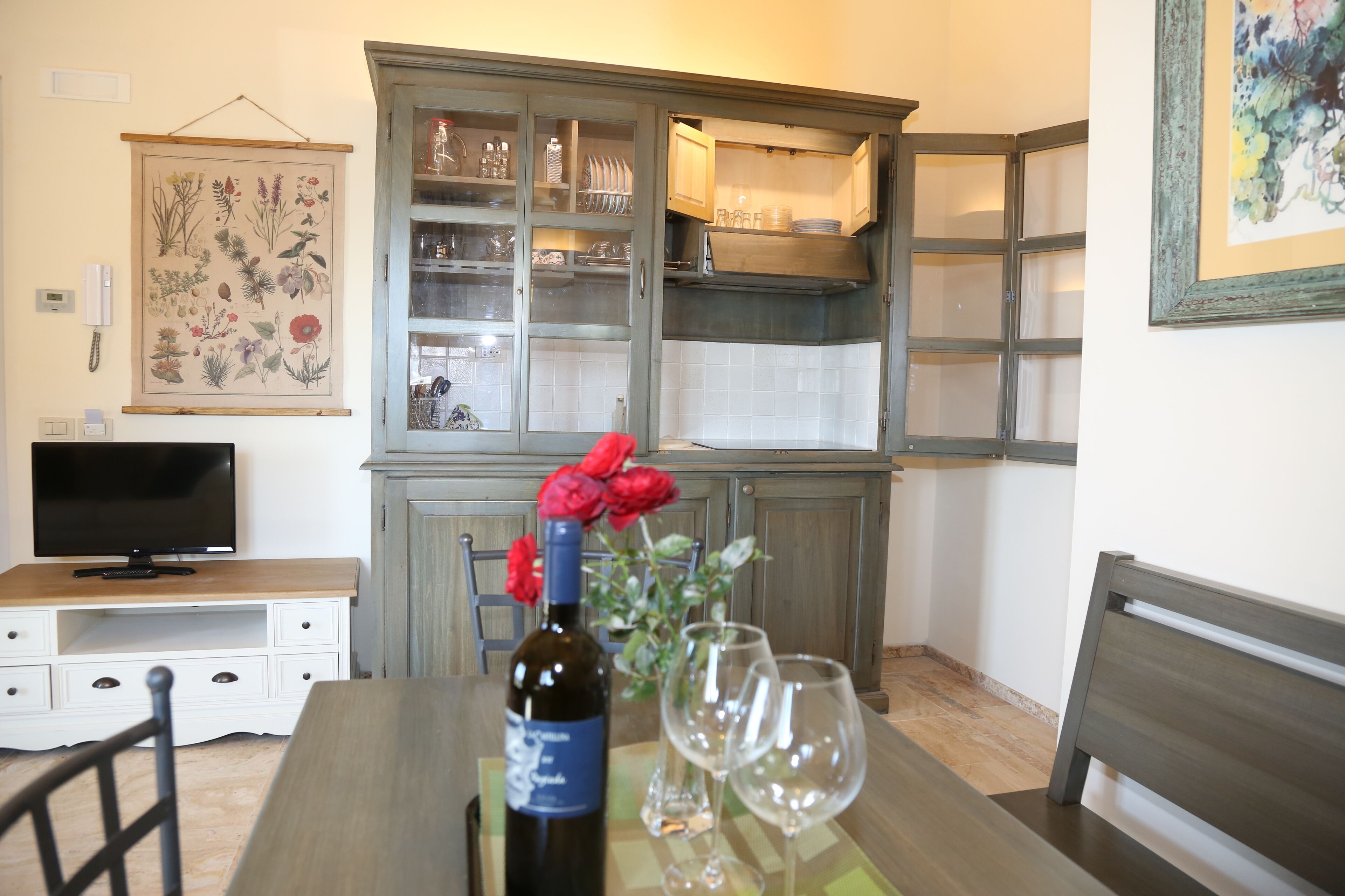 A dining area with a table set for two, a bottle of wine, and two wine glasses. A cabinet with glass doors is in the background, and a television is on a stand to the left.
