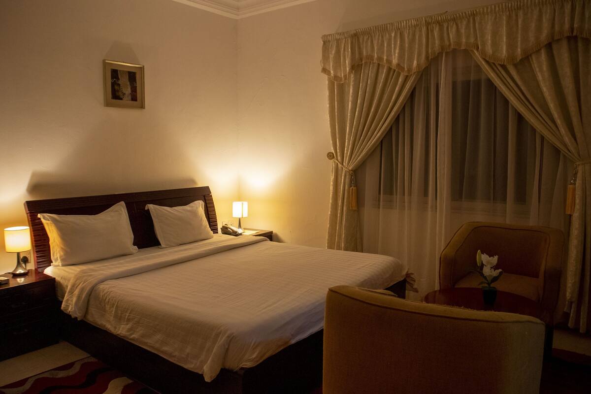 Une chambre d’hôtel avec un grand lit, deux tables de chevet avec des lampes, une chaise et une fenêtre avec des rideaux.