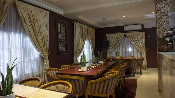 A dining area with a long wooden table and striped chairs, large windows with curtains, and a potted plant on the table.