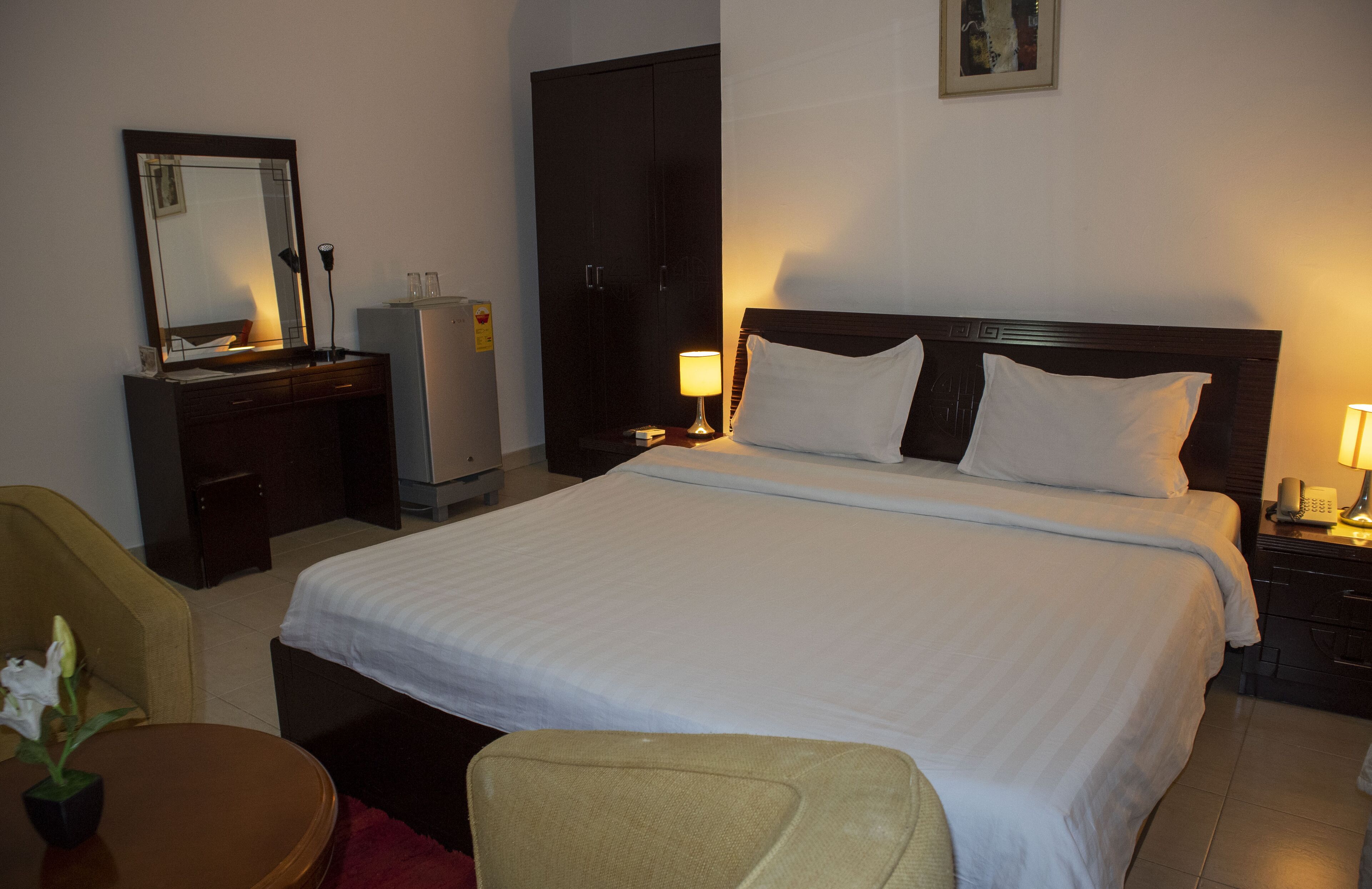 A hotel room with a large bed, a desk with a lamp, a wardrobe, and a chair.