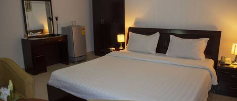 A hotel room with a large bed, a desk with a lamp, a wardrobe, and a chair.