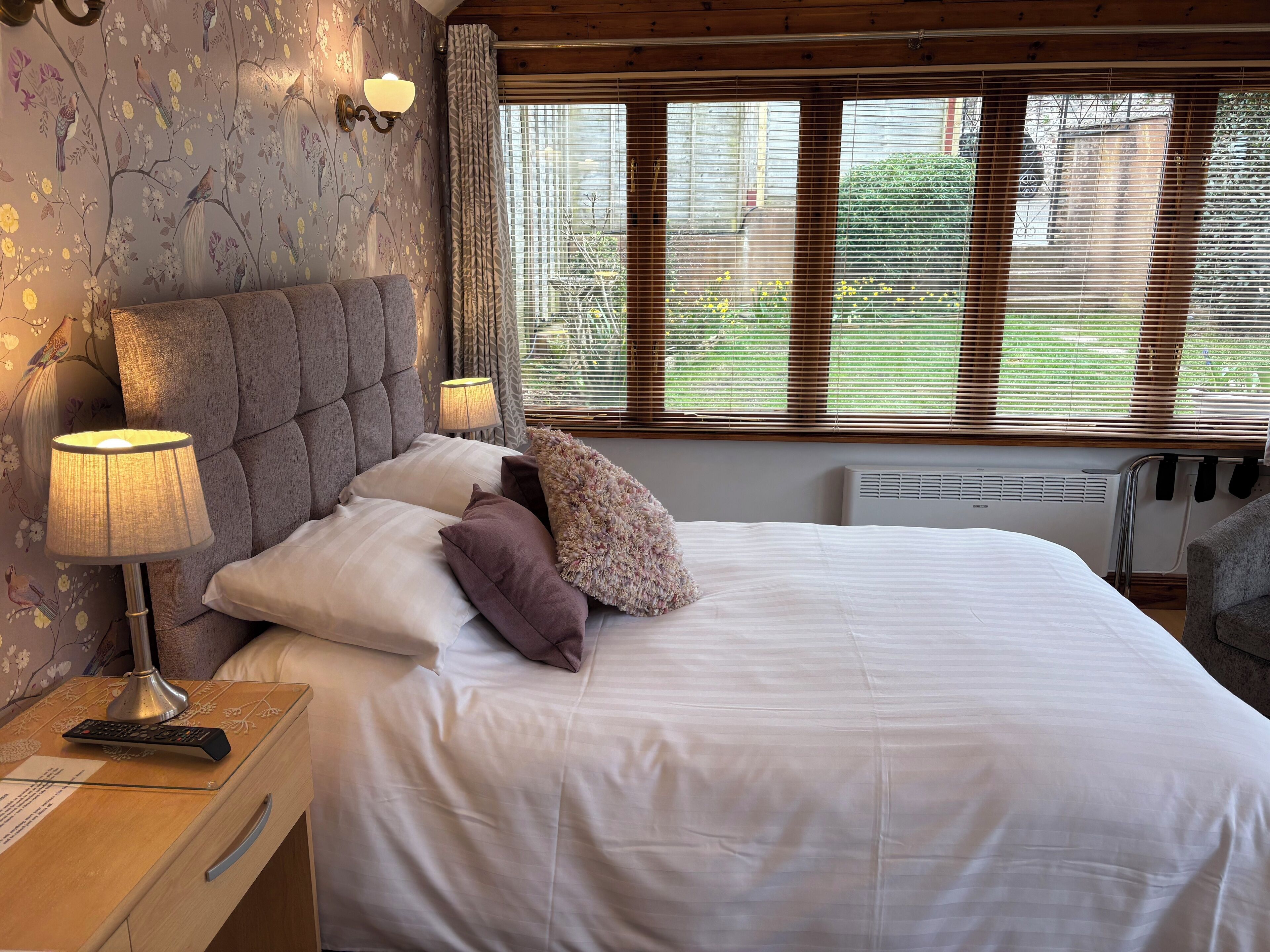 A bedroom with a large bed, a nightstand with a lamp, a chair, and a window with blinds.