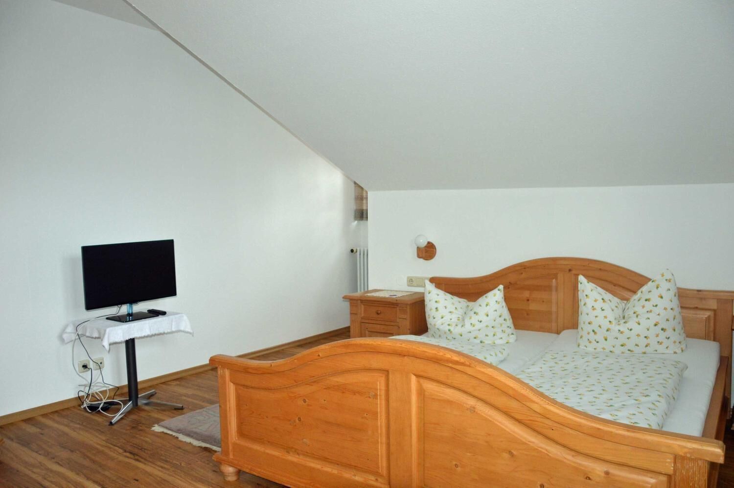 Un dormitorio con cama de madera, televisor sobre un mueble bajo, mesita de noche y otra mesita auxiliar.