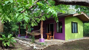 Ein kleines, eingeschossiges Haus mit violetten Wänden und einem grünen Dach, umgeben von üppiger Vegetation.