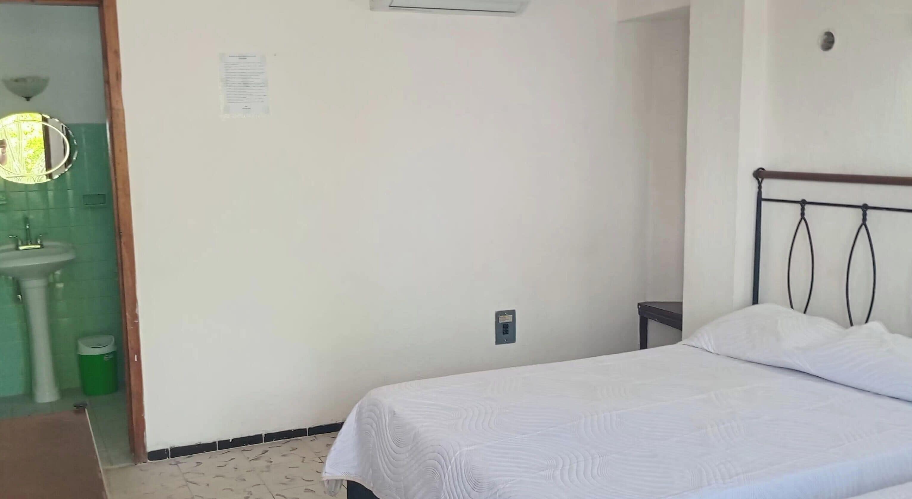 A bedroom with a bed, a nightstand, a wall-mounted light fixture, a green trash bin, and a white sink.