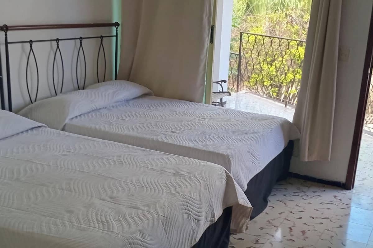 Een kamer met twee bedden, een balkon met metalen hekwerk en een raam met gordijnen.