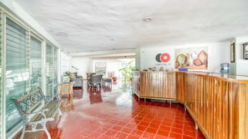 Un hall espacioso con suelo de baldosas rojas, un mostrador de recepción de madera y un amplio ventanal con persianas.