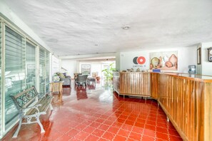A spacious lobby with a red tiled floor, a wooden reception desk, and a large window with blinds. - Hotel San Carlos (Tizimín)