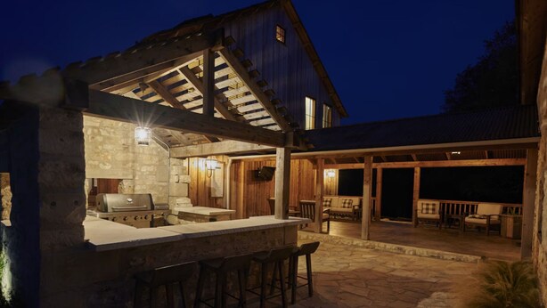 A covered outdoor kitchen with stone walls, a grill, and bar stools. - Entire Home at Melissa's Farmstead (Fredericksburg)