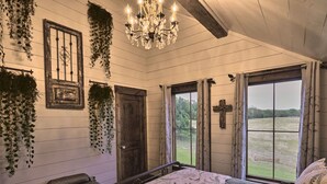 A bedroom with a bed, a chandelier, a window with curtains, and a door. - Entire Home at Melissa's Farmstead (Fredericksburg)