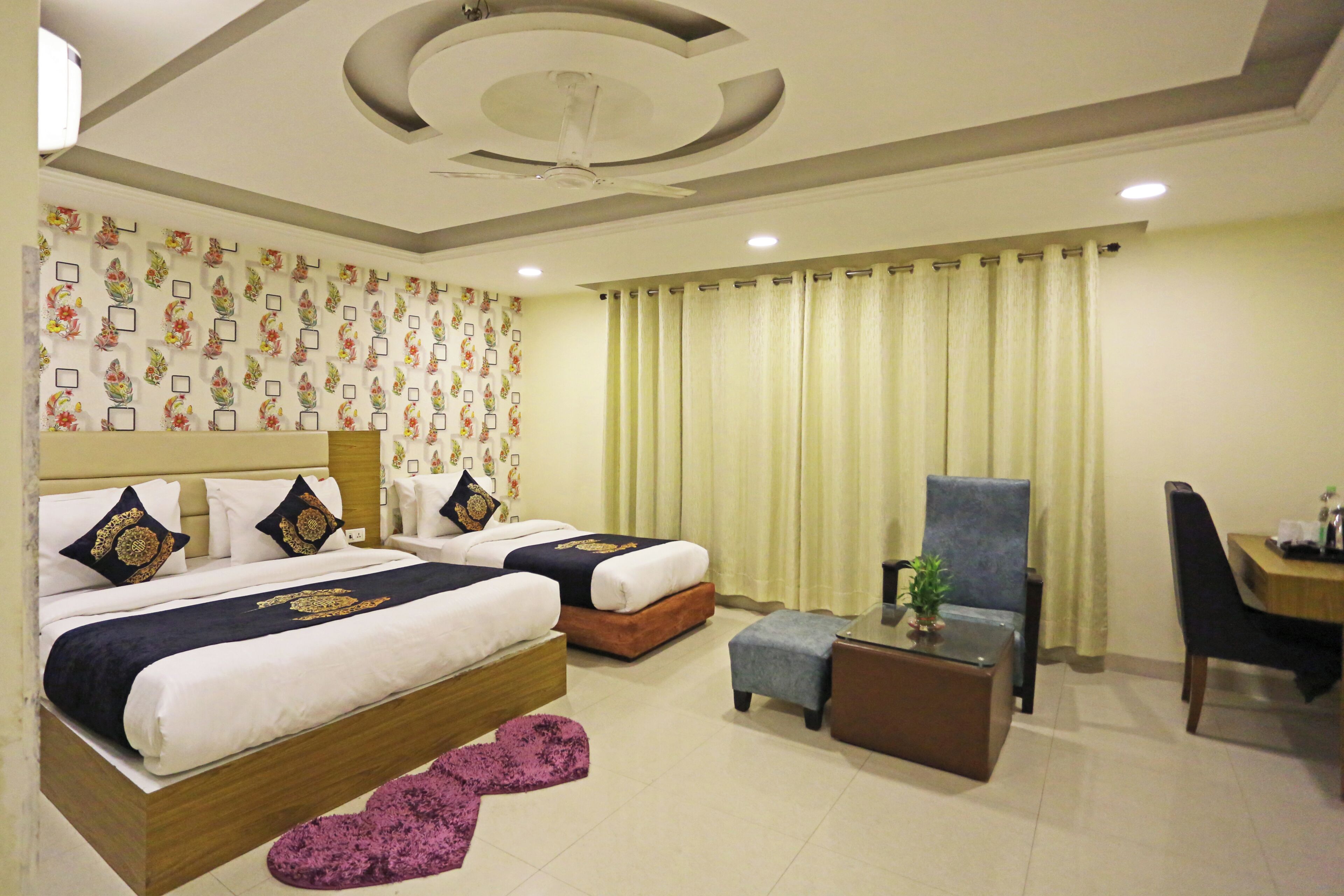 A hotel room with two beds, a desk, a chair, a small table, a ceiling fan, and a patterned wall.