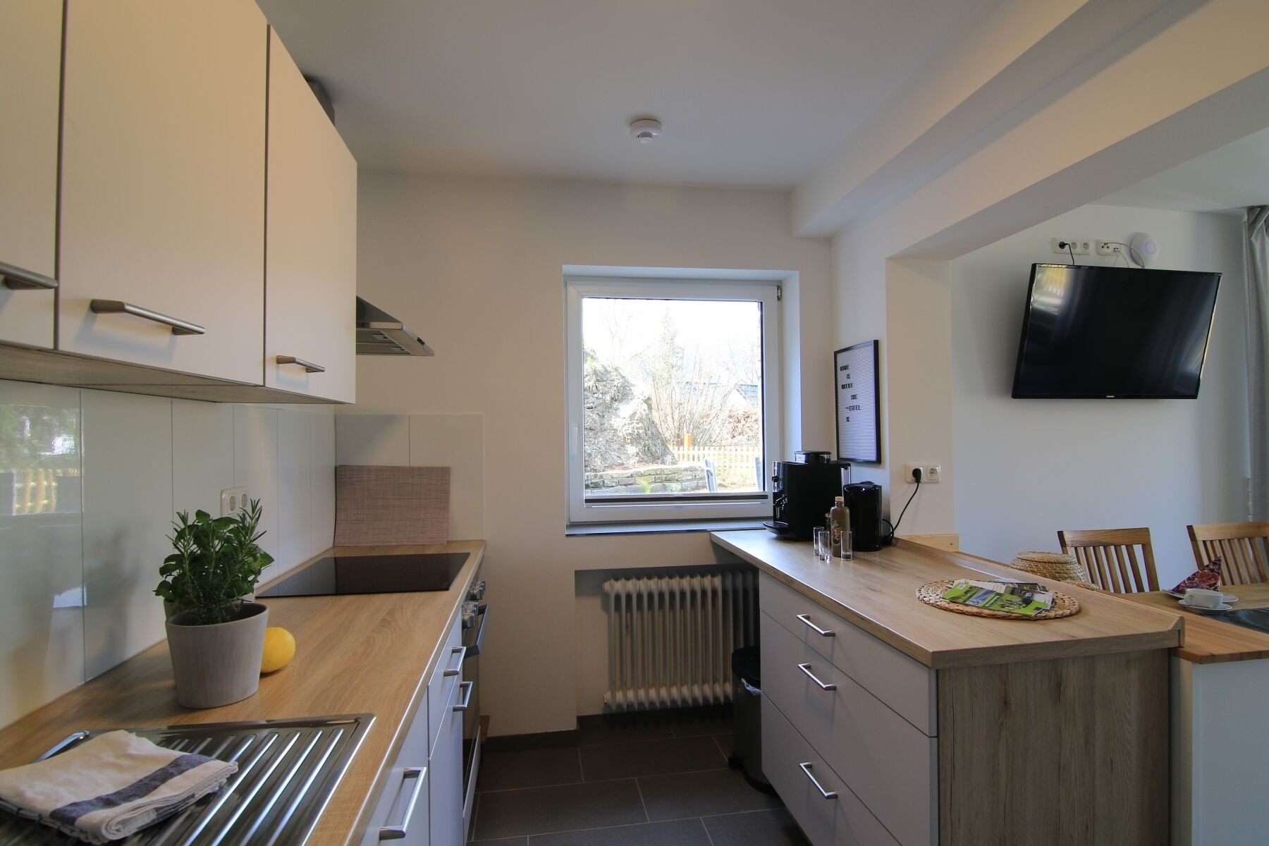 Una cucina moderna con mobili bianchi, un piano di lavoro in legno, una finestra con vista, un piano cottura integrato e un televisore a schermo piatto montato a parete.