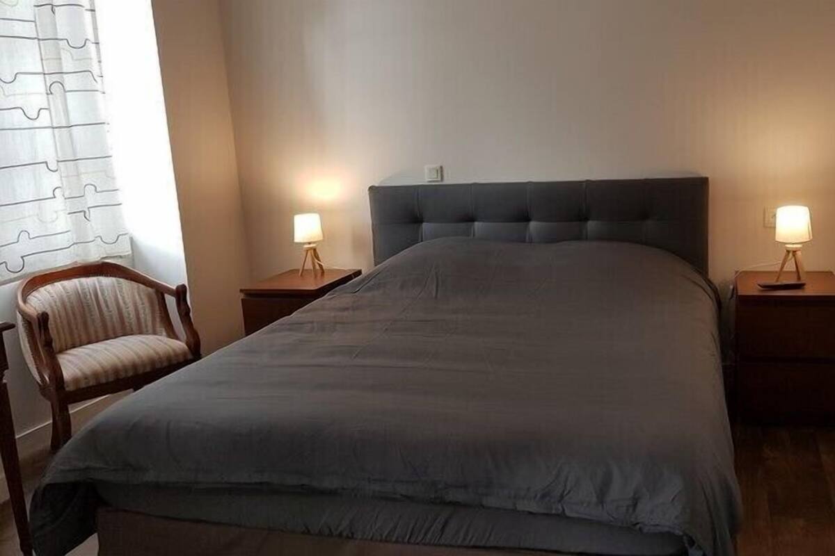 A bedroom with a large bed, two bedside tables with lamps, a wooden chair, and a window with curtains.