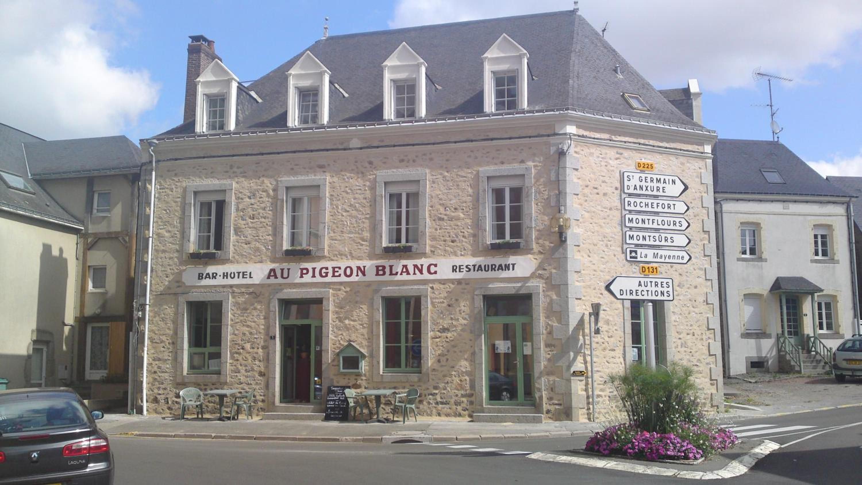 A stone building with a sign for 'BAR-HOTEL AU PIGEON BLANC RESTAURANT' and directional signs for nearby locations.