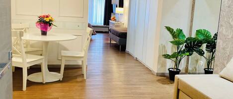 A modern hotel room with a wooden floor, a white dining area with a table and chairs, a sofa, and a large mirror.
