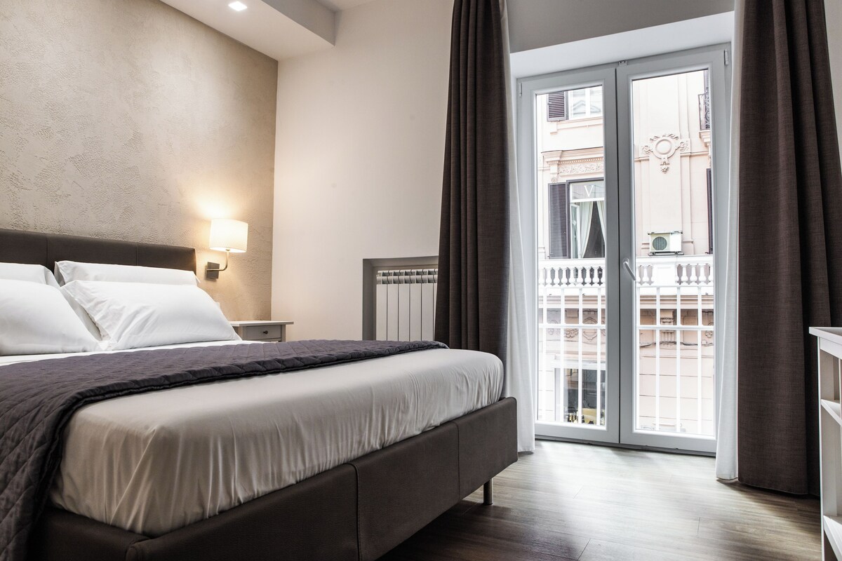 A modern hotel room with a large bed, a nightstand, a lamp, and a view of a balcony with a railing.