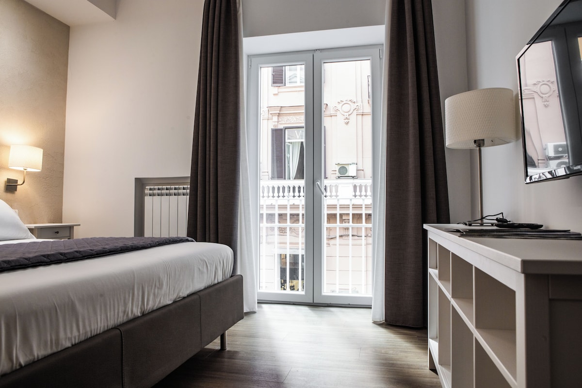 Habitació d'hotel amb llit, una taula de nit, una làmpada, un armari i vista a l'exterior.