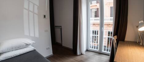 A hotel room with a bed, a desk with a lamp, a radiator, and a view of a building through a door.