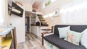 A compact living space with a sofa, a TV mounted on the wall, a kitchen area, and a staircase. - Tiny House Siesta- perfect tiny house for two! (Sarasota)