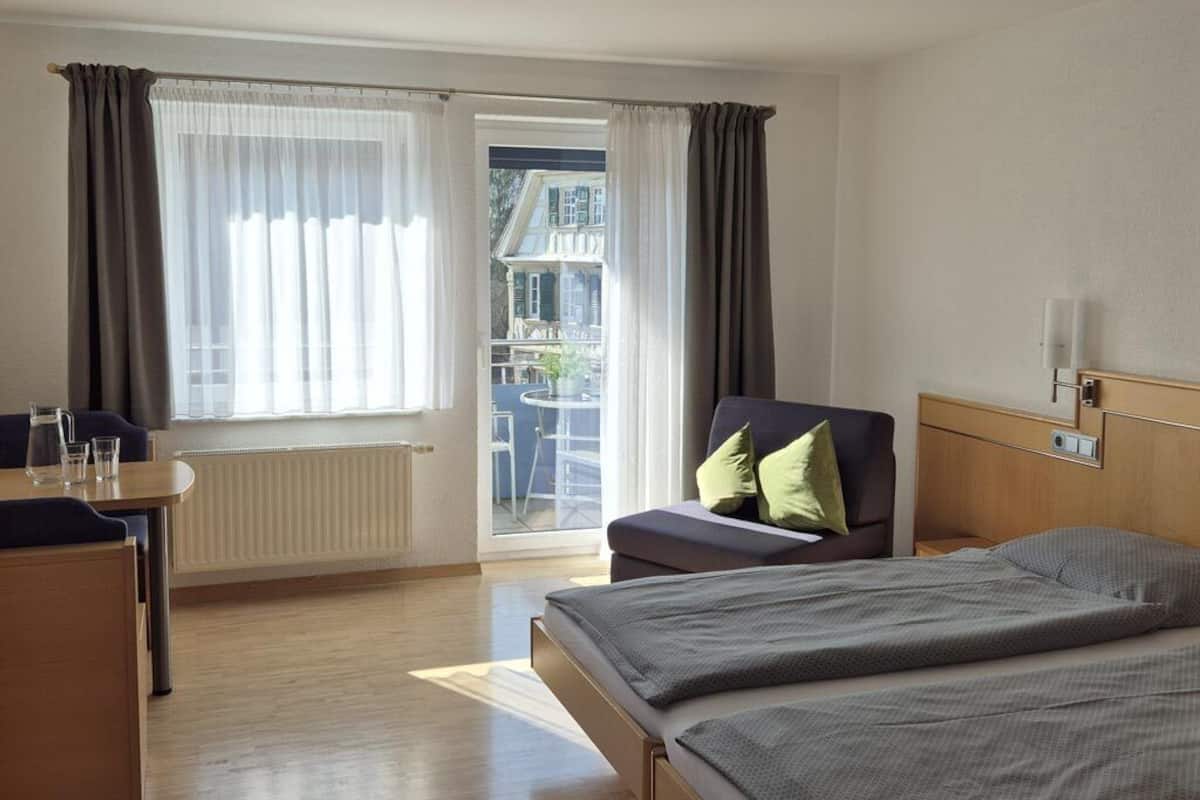A hotel room with a bed, a chair, a table, and a view of the outside.