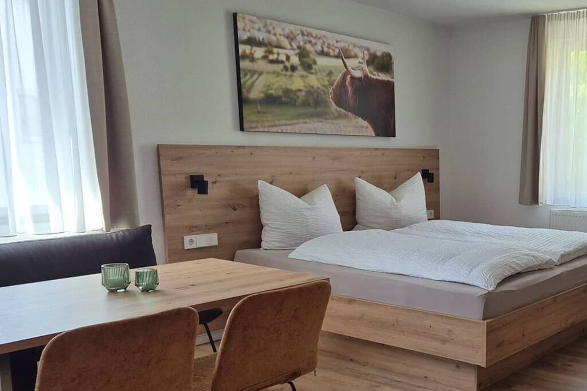 A bedroom with a wooden bed, white pillows, a wooden headboard, a dining table with chairs, and a painting on the wall.