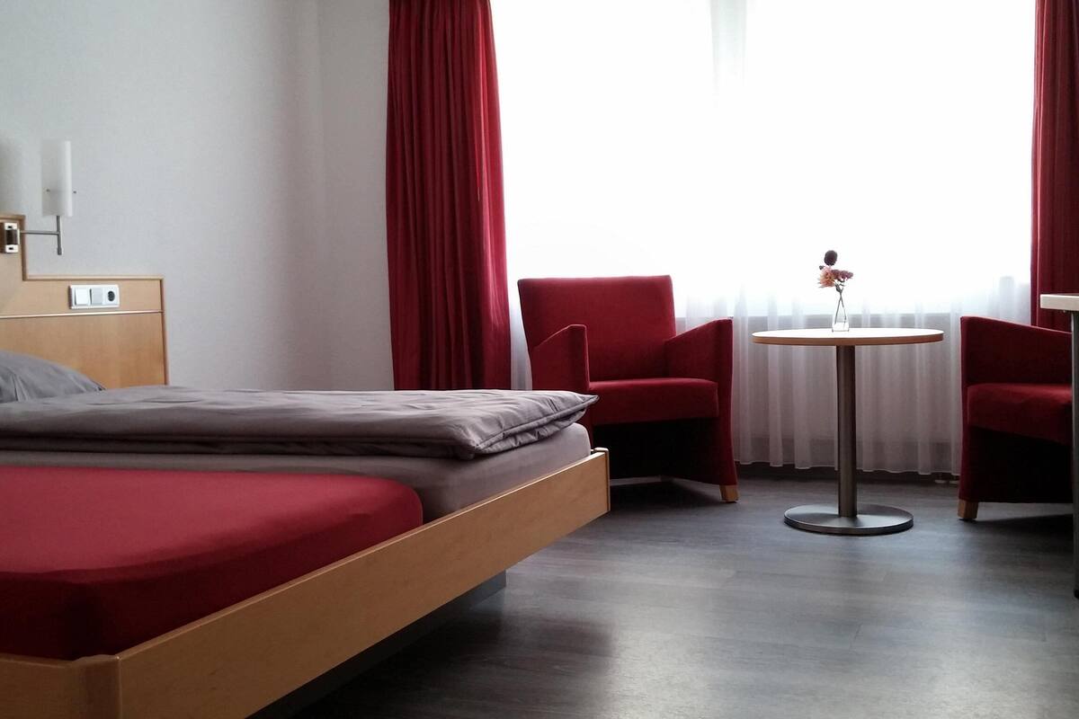 A hotel room with a bed, a small table, two red chairs, and a window with red curtains.
