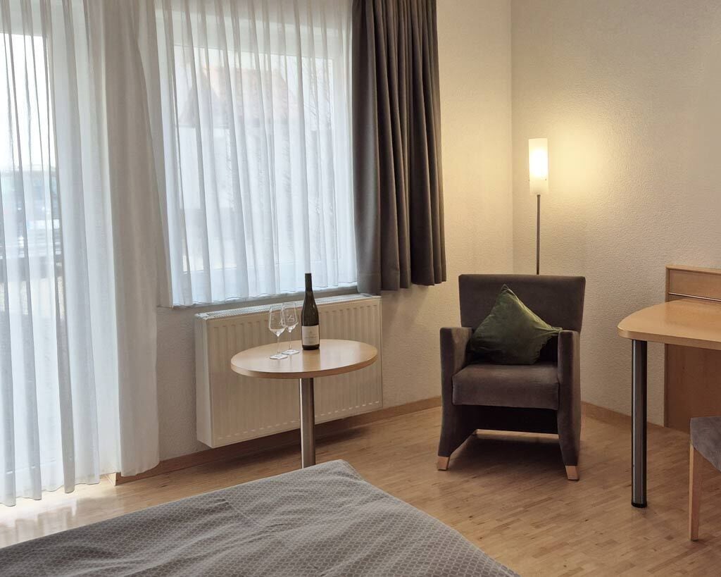 Een hotelkamer met een bed, een stoel, een klein tafeltje met een fles, een lamp en een raam met gordijnen.