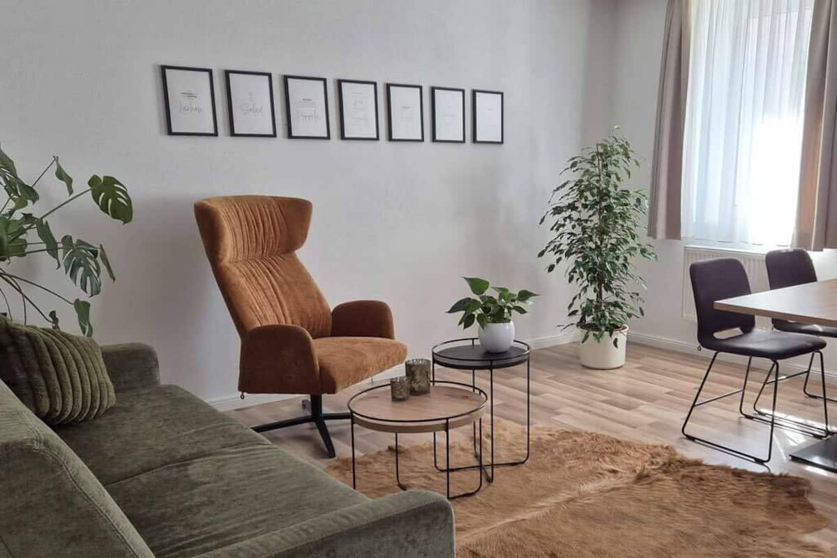 A living room with a green sofa, a brown armchair, a coffee table, a dining table with chairs, and a potted plant.