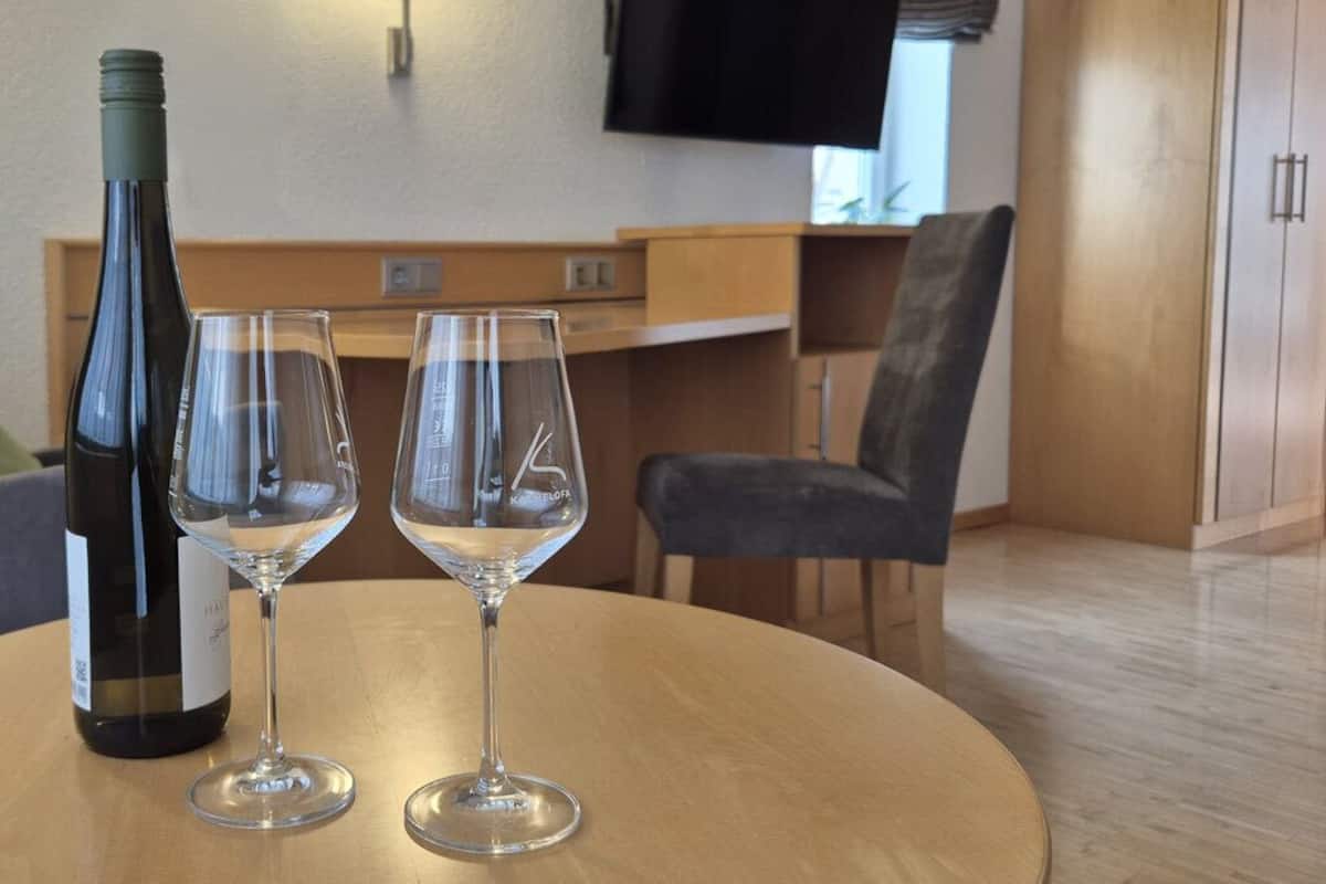 A room with a round table, two wine glasses, a bottle of wine, a flat-screen TV mounted on the wall, a wooden cabinet, and a chair.