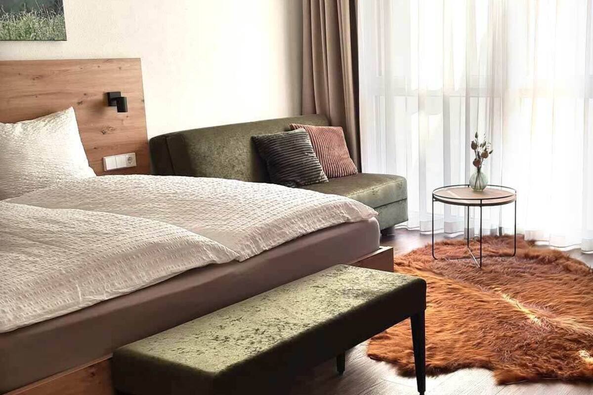 A bedroom with a bed, bench, sofa, and a small table with a vase.