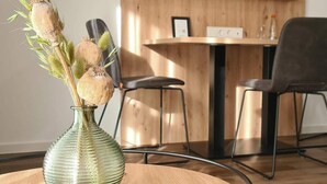 A room with a round wooden table, a green vase with dried flowers, and a TV mounted on the wall.