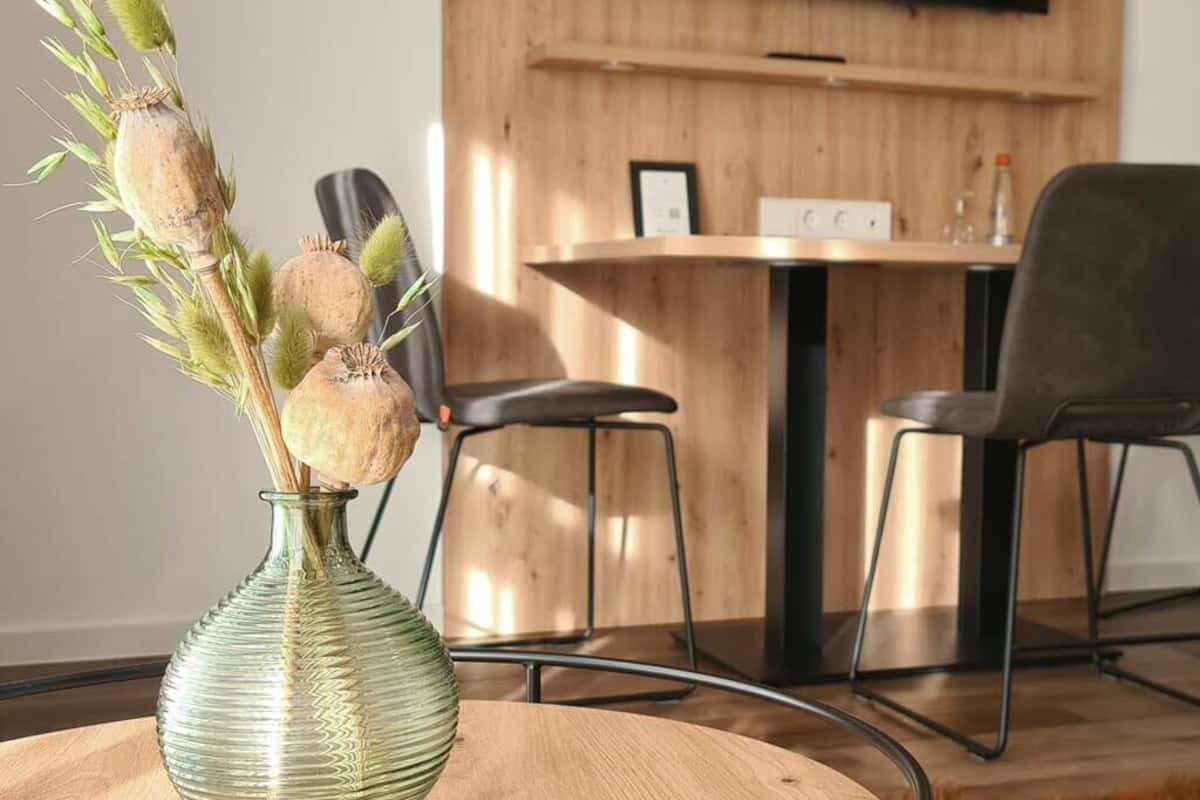 A room with a round wooden table, a green vase with dried flowers, and a TV mounted on the wall.