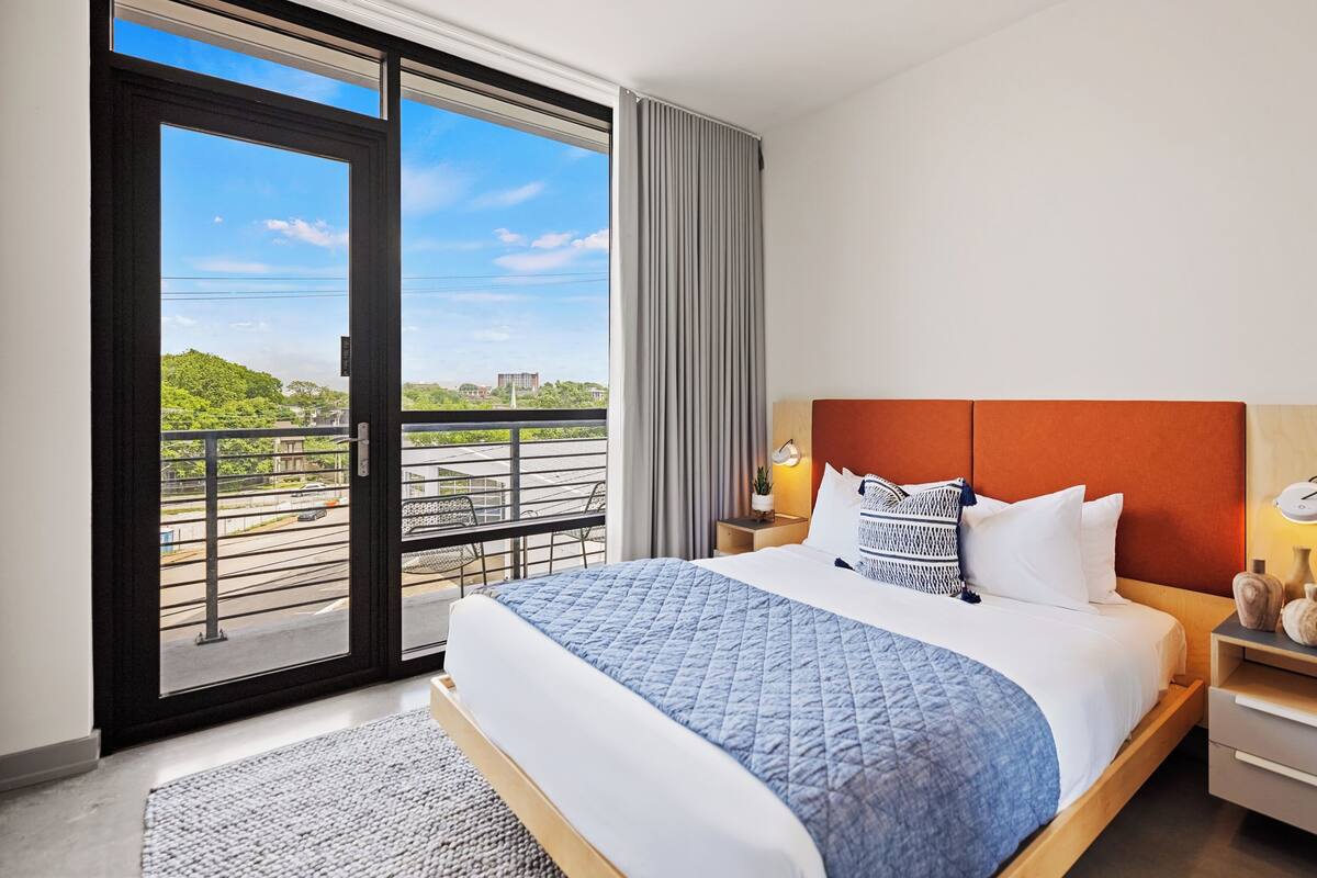 Une chambre d’hôtel avec un grand lit, une table de chevet, une lampe fixée au mur et une vue sur un paysage verdoyant, visible à travers une porte coulissante.
