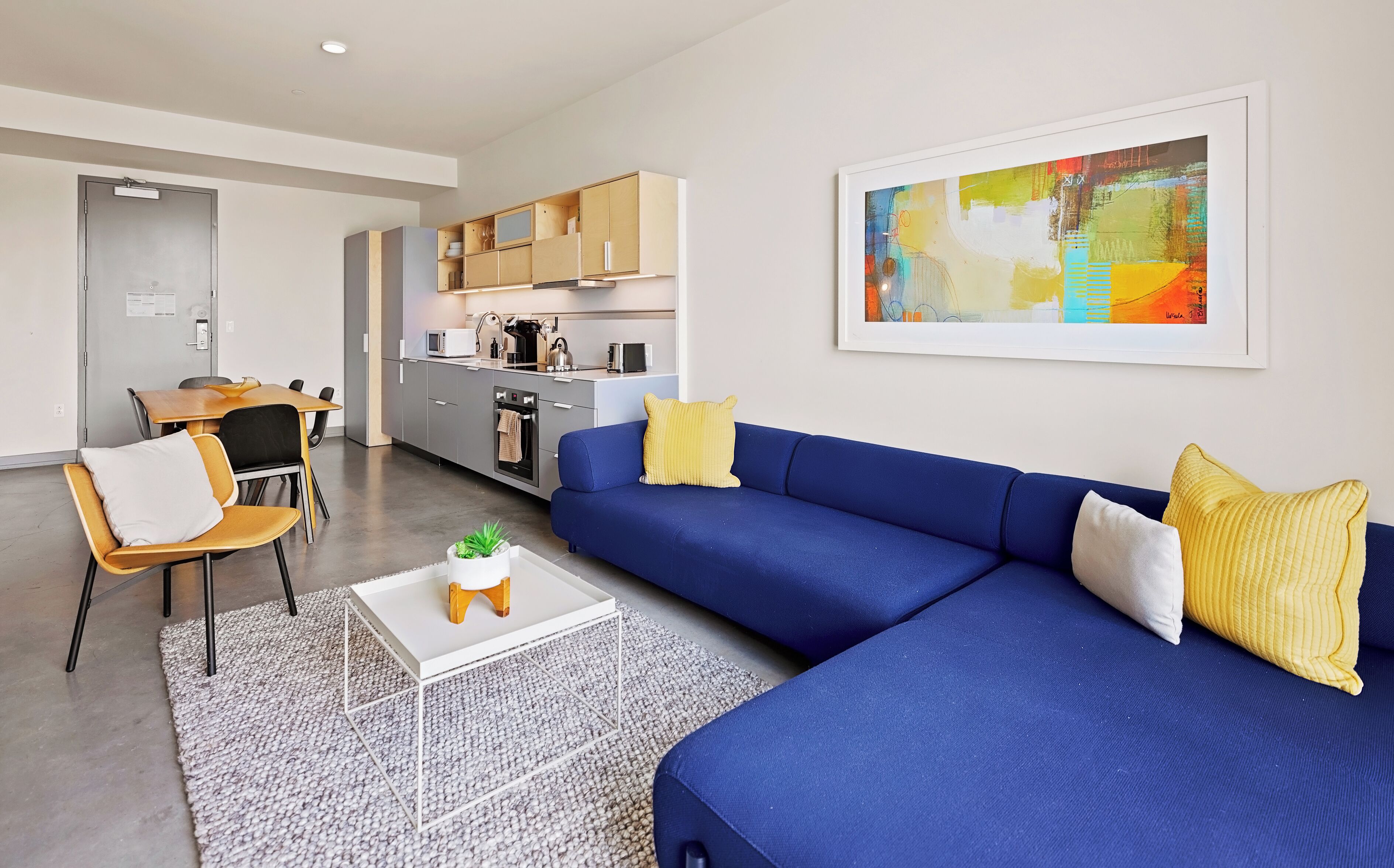 Ein modernes Wohnzimmer mit einem blauen Sofa, gelben Kissen, einem weißen Couchtisch mit einer Pflanze und einer Küche im Hintergrund.