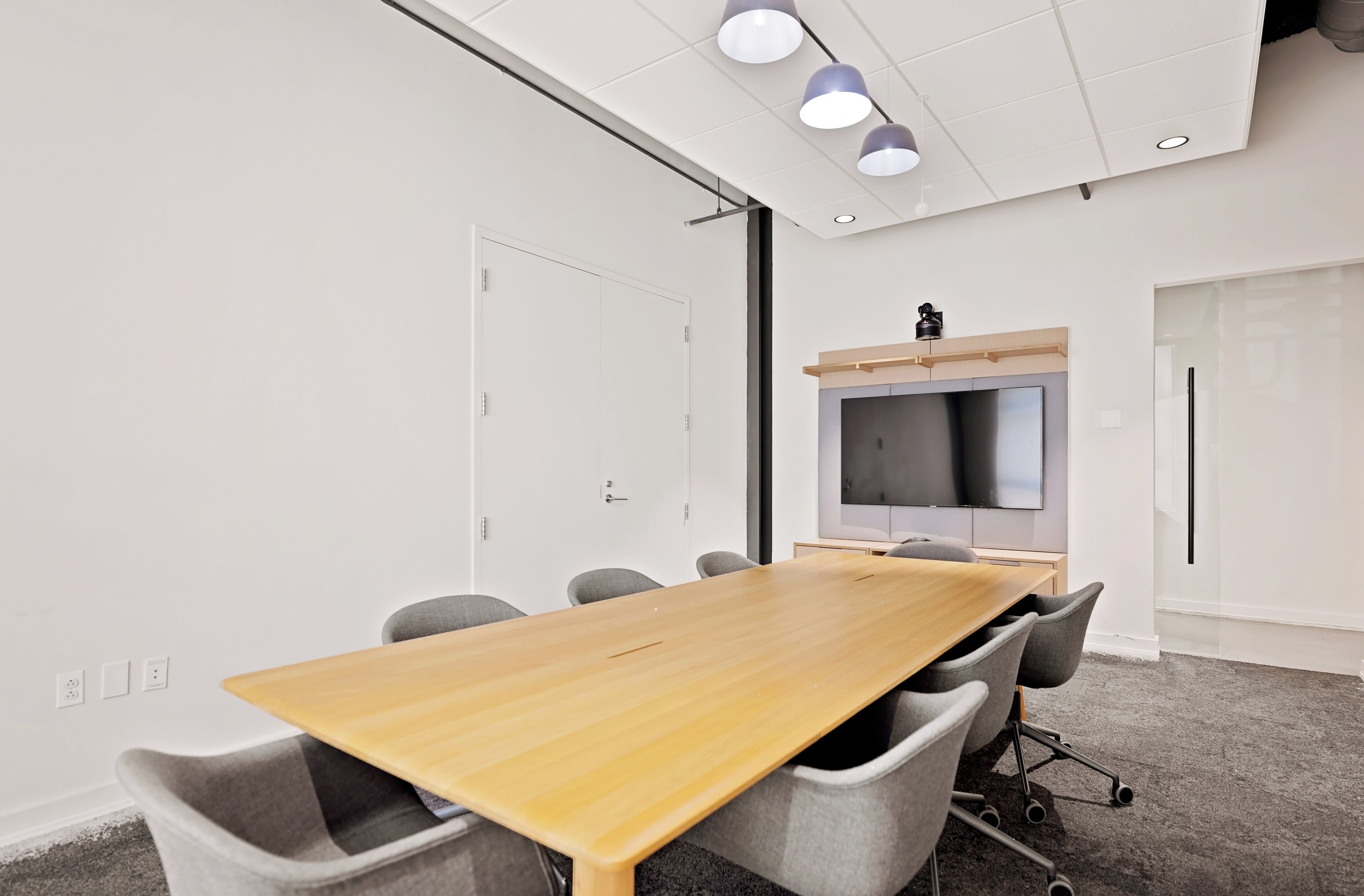 Een vergaderruimte met een lange houten tafel, grijze stoelen, een flatscreen televisie aan de muur en in het plafond verzonken verlichting.