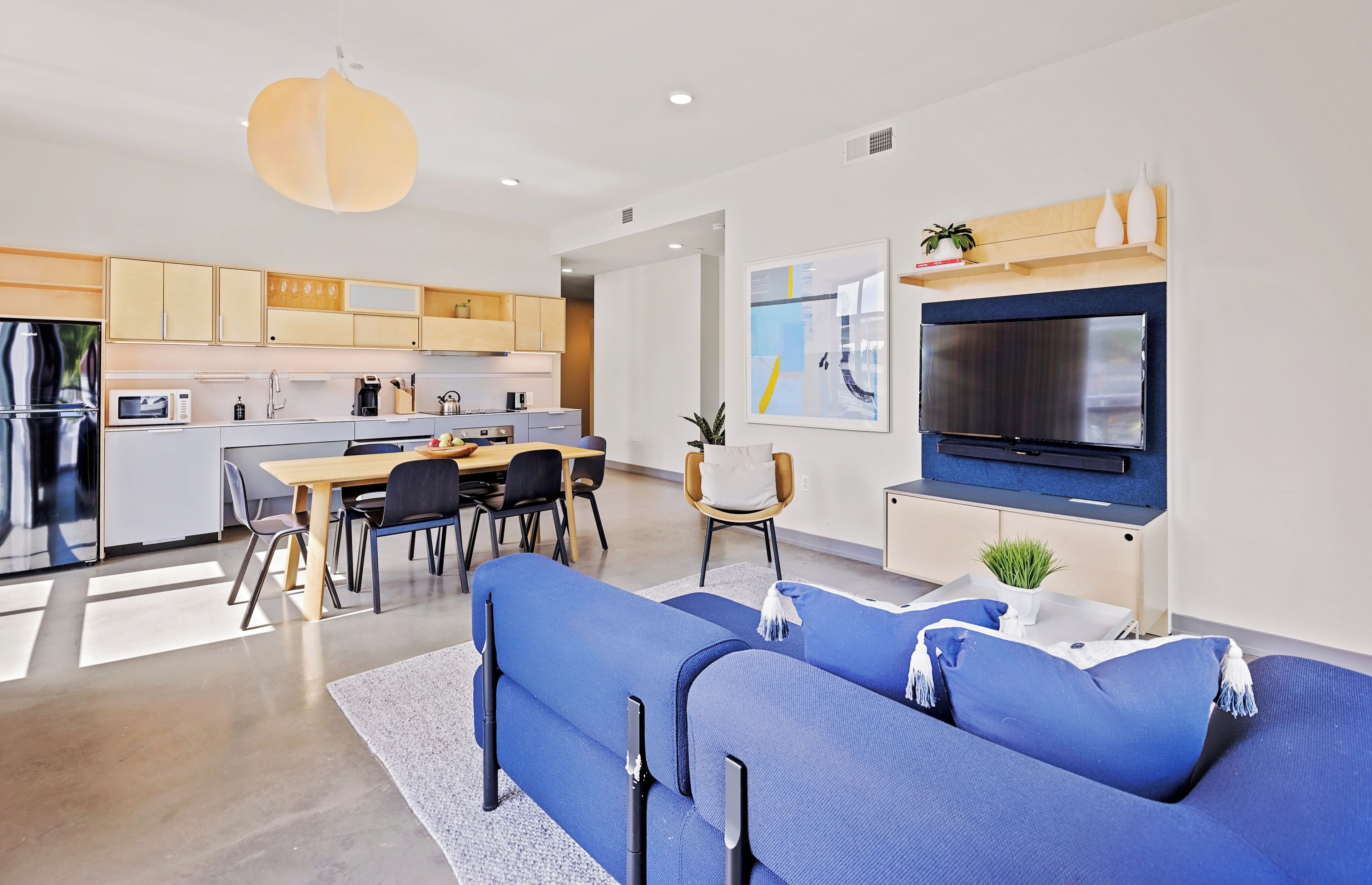 A modern kitchen with built-in appliances, a dining area with a table and chairs, a large TV, and a blue sofa.
